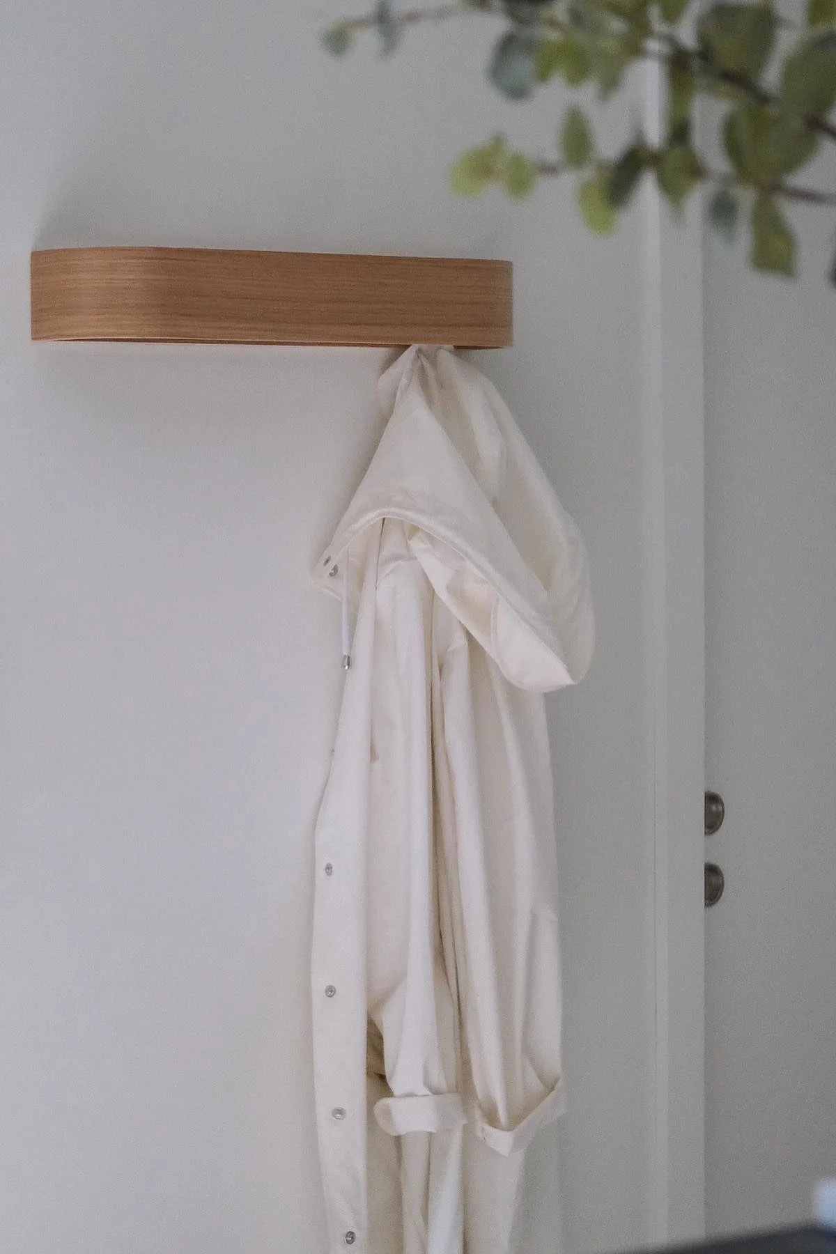 A light-colored hooded jacket hanging on a minimalist light wood wall hook, against a white wall with blurred greenery.