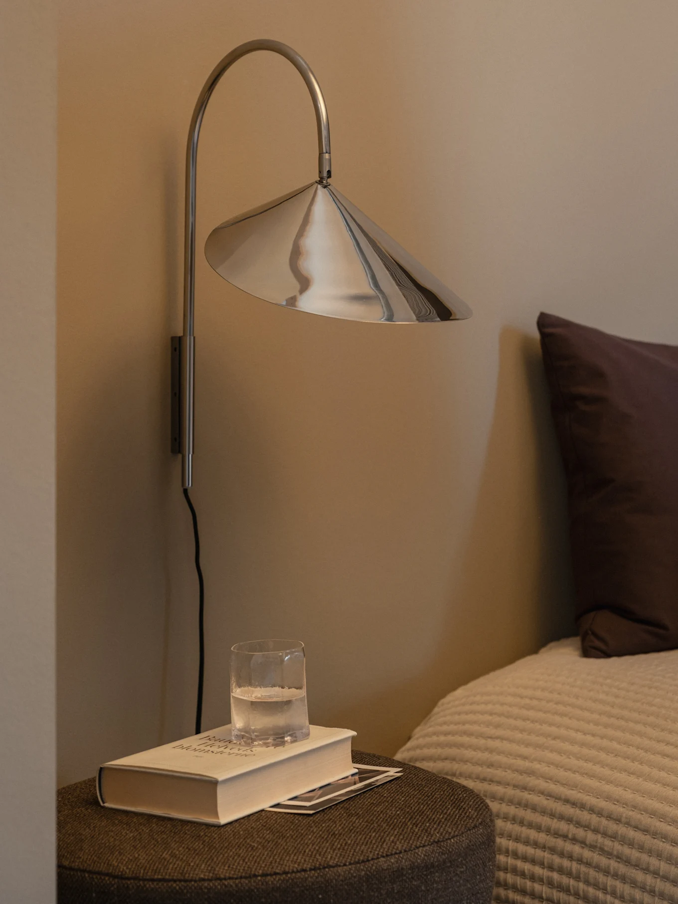 A modern silver wall lamp with a conical shade above a book and a glass of water on a bedside table.