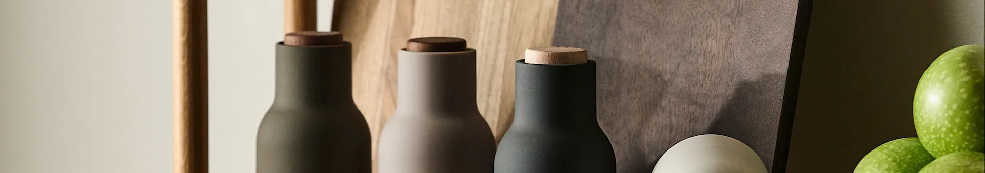 Three modern grinders in dark green, beige, and black with wooden tops, against a wood background with green apples.