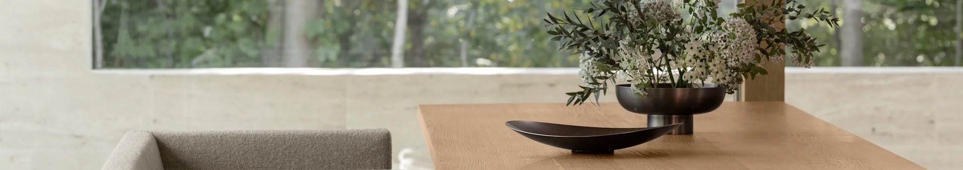 Modern wooden dining table with a decorative dark bowl and a vase of white and green flowers, with a blurry green outdoor view through a large window.