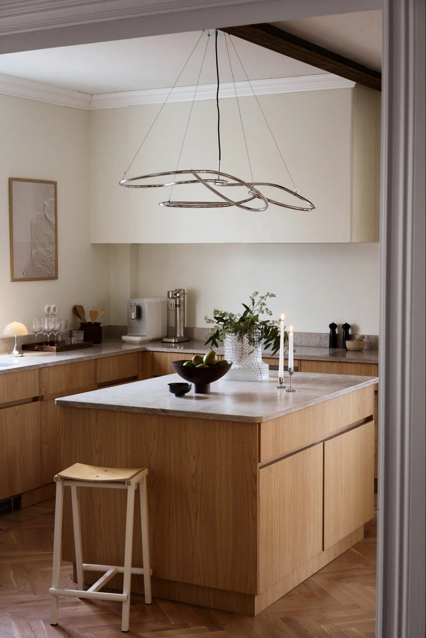 A modern kitchen with light wood cabinets, marble counters, a large island, and a sculptural chrome chandelier.