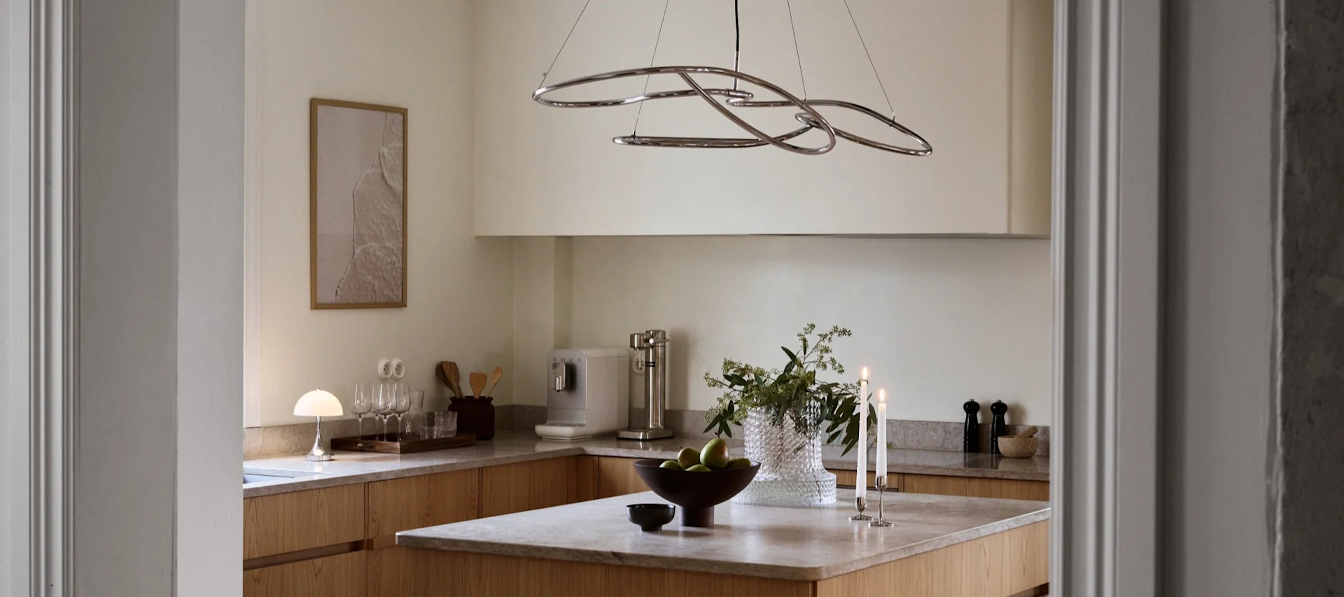 A modern kitchen with light wood cabinets, an island featuring green apples, lit candles, and a sculptural chrome chandelier.