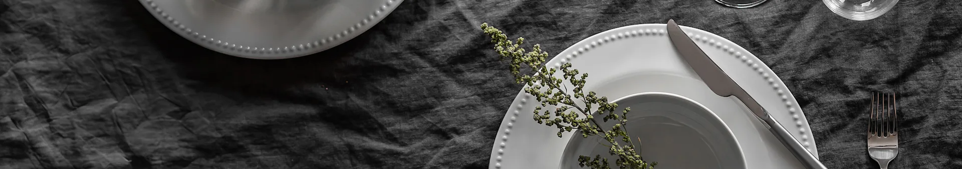 Elegant table setting with white beaded plates, silverware, a glass, and a green sprig on a dark grey linen tablecloth.