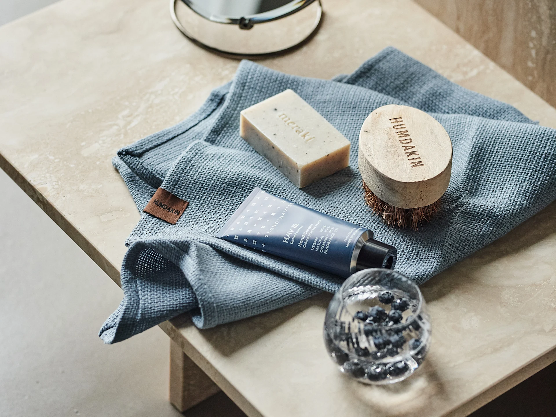 Blue towel, bar of soap, wooden hand brush, blue hand cream tube, and a glass with blueberries on a light countertop.