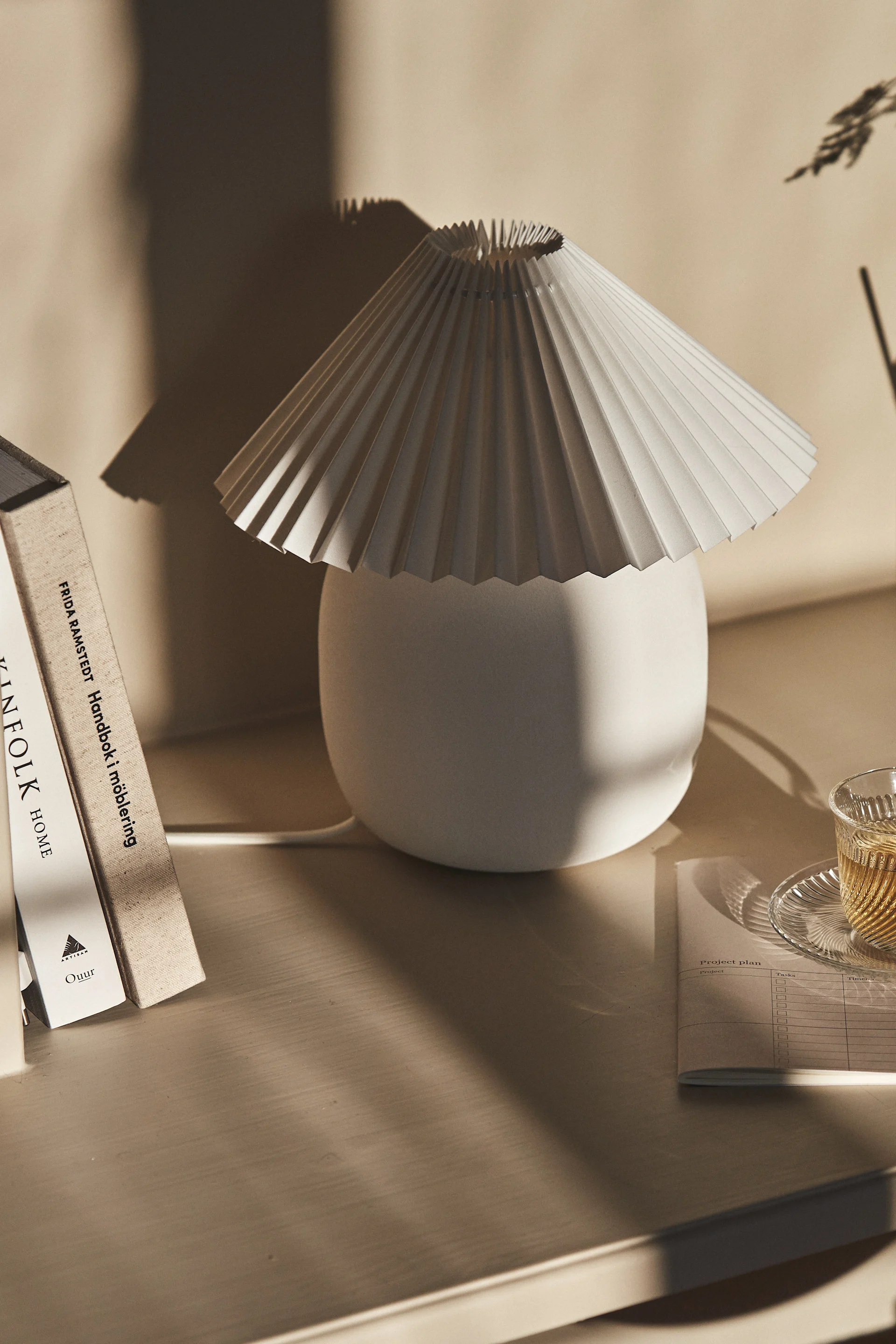 A pleated white table lamp next to books and a cup of tea on a light wooden desk, with soft shadows.