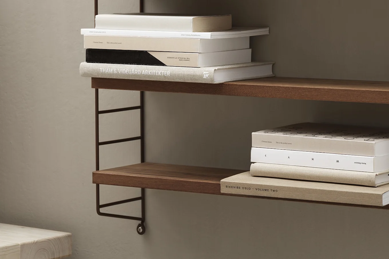 Two wooden shelves on a dark metal string system against a light brown wall, holding stacks of books.