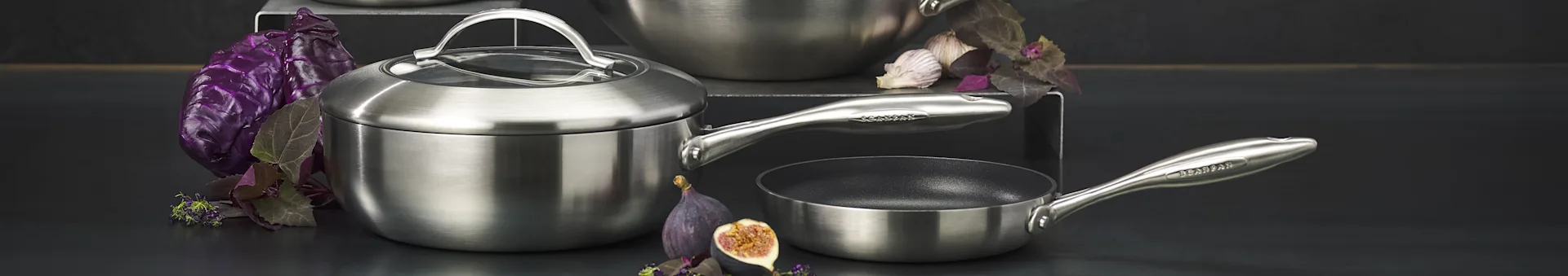 Brushed stainless steel cookware set: lidded pot, frying pan, surrounded by purple cabbage, garlic, and figs.