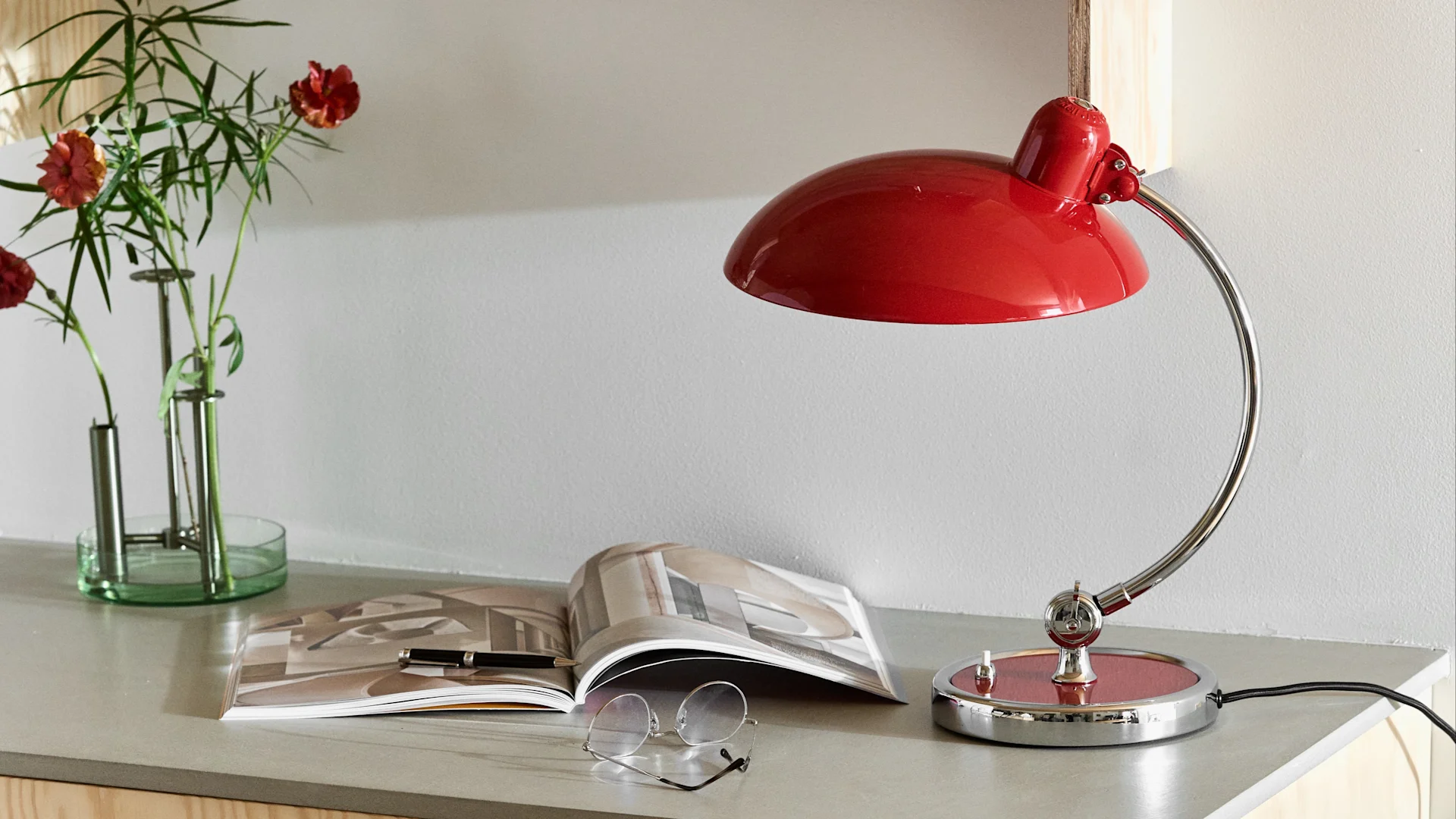A desk scene with a red curved desk lamp, an open magazine, a pen, eyeglasses, and a vase of red flowers and greenery.