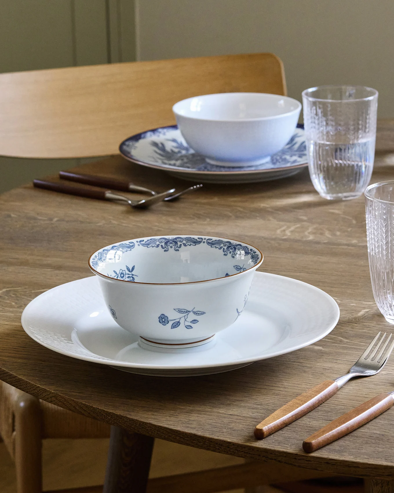 Dining table set for two with white and blue patterned bowls on white plates, wooden handled cutlery, and water glasses.
