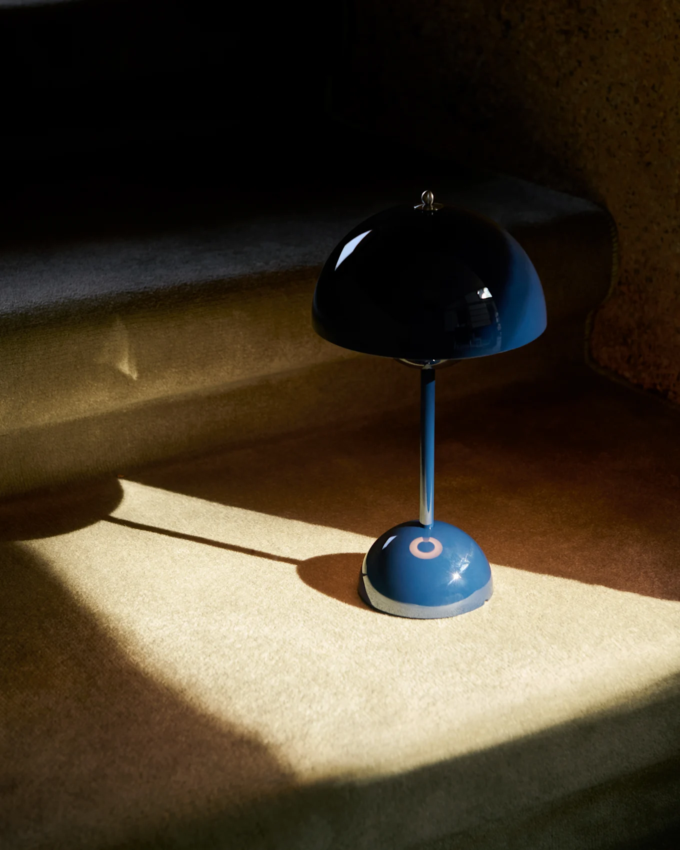 A blue mushroom-shaped table lamp with a reflective shade sits on a carpeted surface, partially illuminated by sunlight.