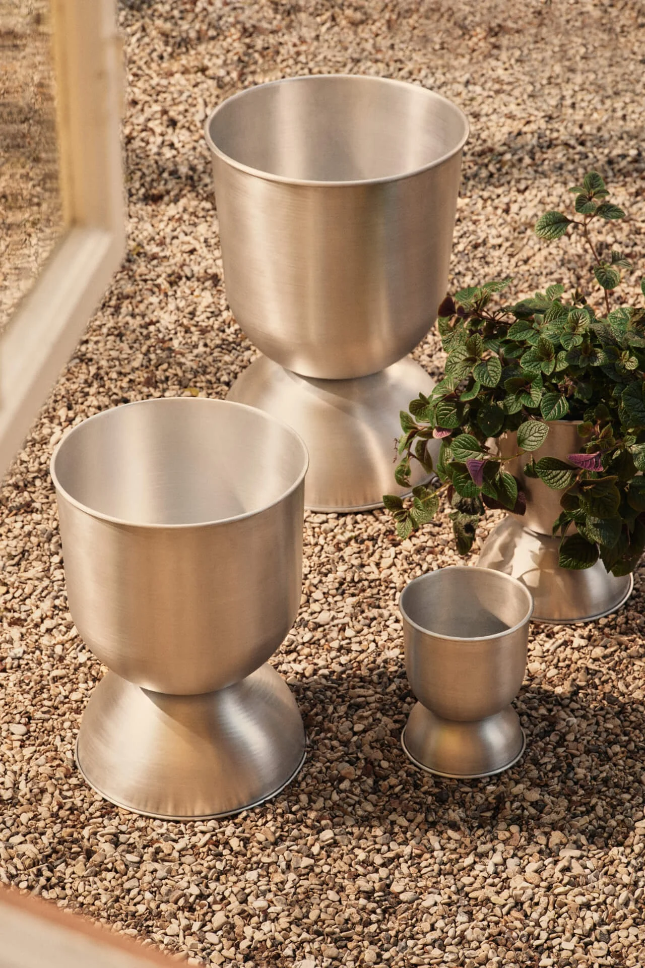 Three brushed metal planters of different sizes on pebbles, one with a green plant.