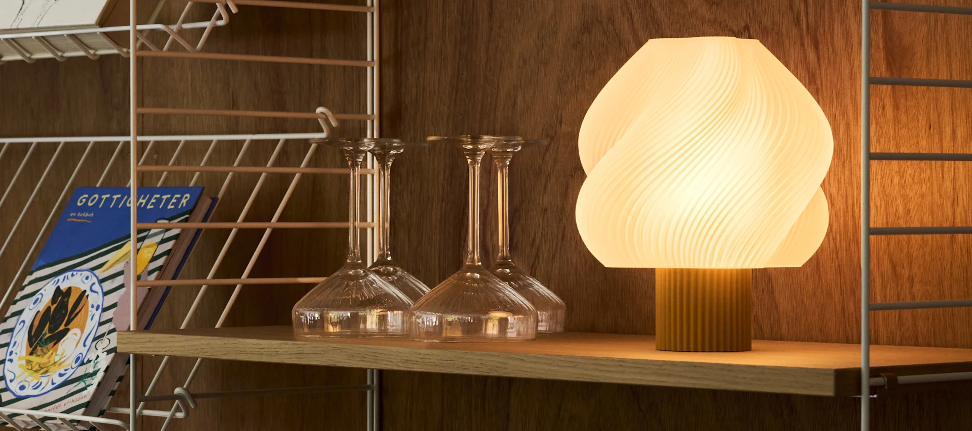 An illuminated, ribbed oval lamp and various clear glasses sit on a wooden bar cart against a warm, textured wall.