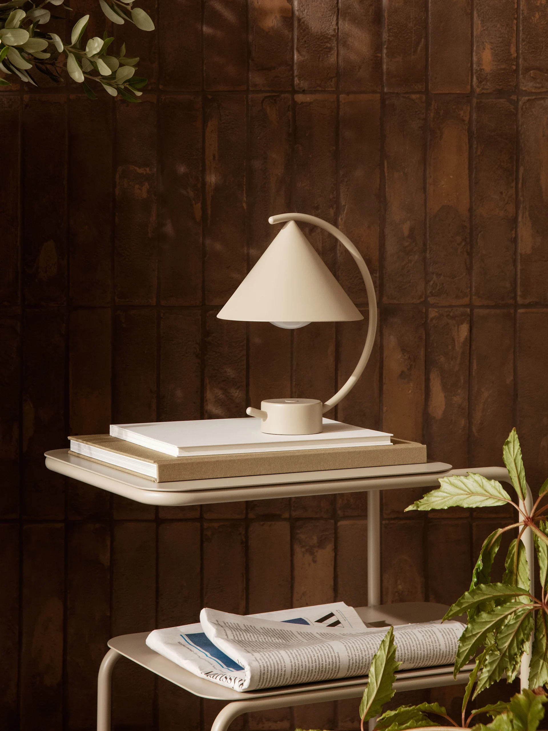 A cream-colored, cone-shaped table lamp sits on books on a light beige side table against a brown tiled wall, with green plants.