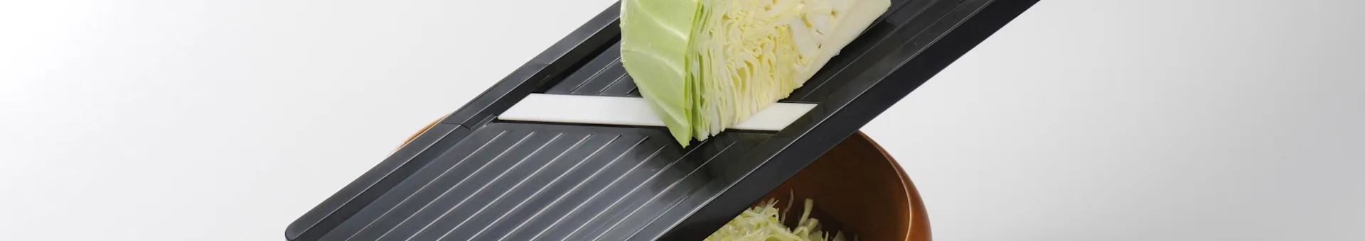 A mandoline slicer with a large wedge of cabbage being shredded into a wooden bowl.