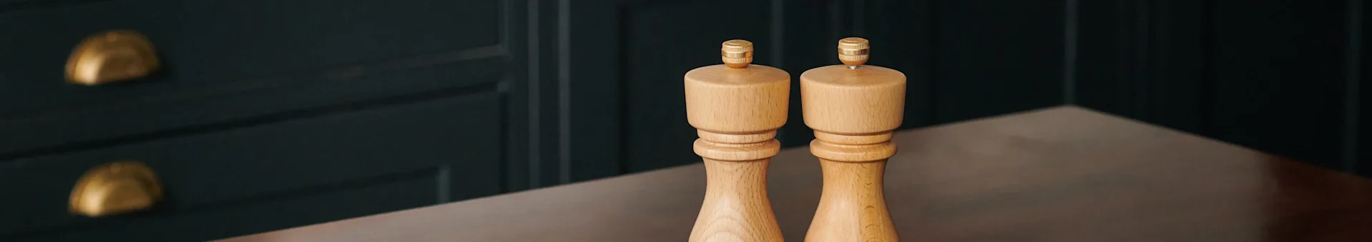 Two wooden salt and pepper grinders with brass tops on a dark table, with a dark cabinet and gold handles in the background.
