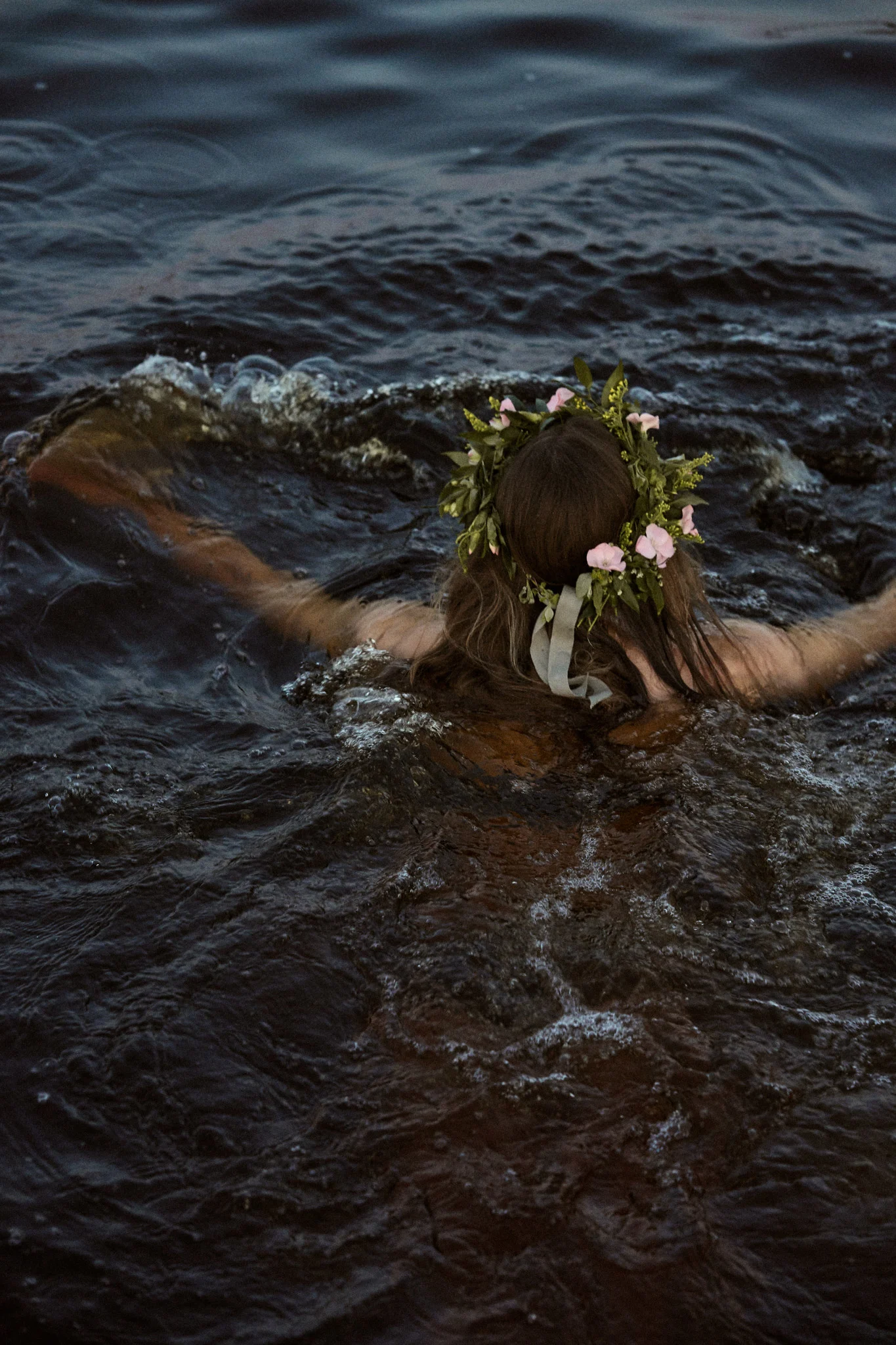 Person with a flower crown swimming in dark water, seen from behind.