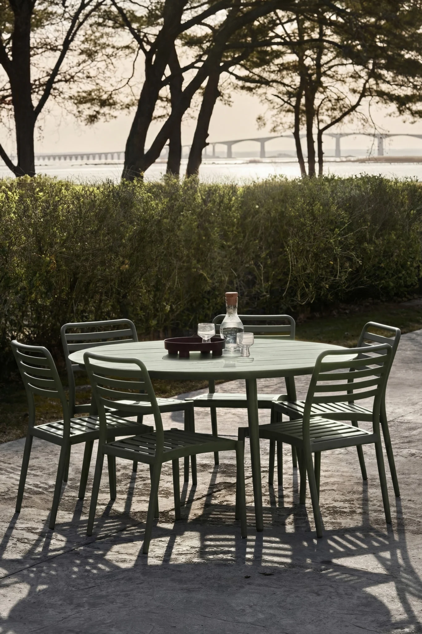 Green outdoor dining set with table, five chairs, water bottle, and glasses on patio. Distant bridge over water.