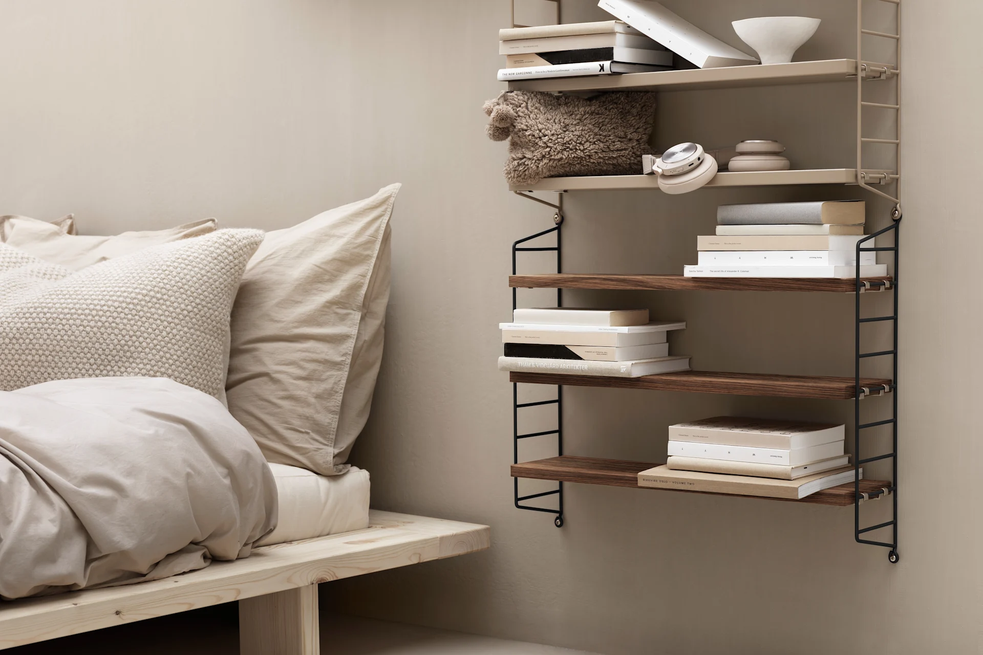 A cozy bedroom with a light wooden bed and beige bedding next to a wall-mounted shelving unit holding books, a pillow, and headphones.