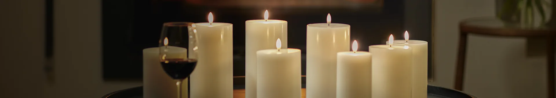 A cozy scene with several lit white pillar candles of varying heights next to a glass of red wine, all set on a dark table.