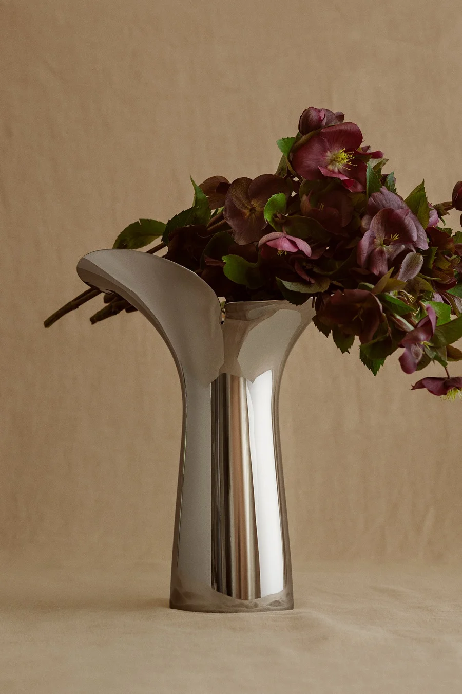 Sleek, reflective silver vase with an organic flared top, holding a bouquet of dark purple flowers and green leaves.