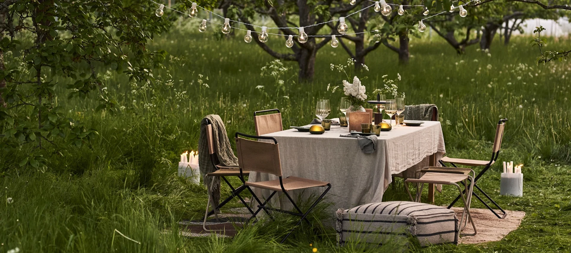 Outdoor dining table set under string lights in a grassy orchard at dusk, with chairs and ambient candles.
