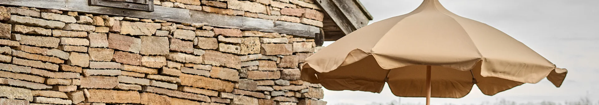 A light brown stone wall on the left, with a wooden beam, next to a beige umbrella on the right.