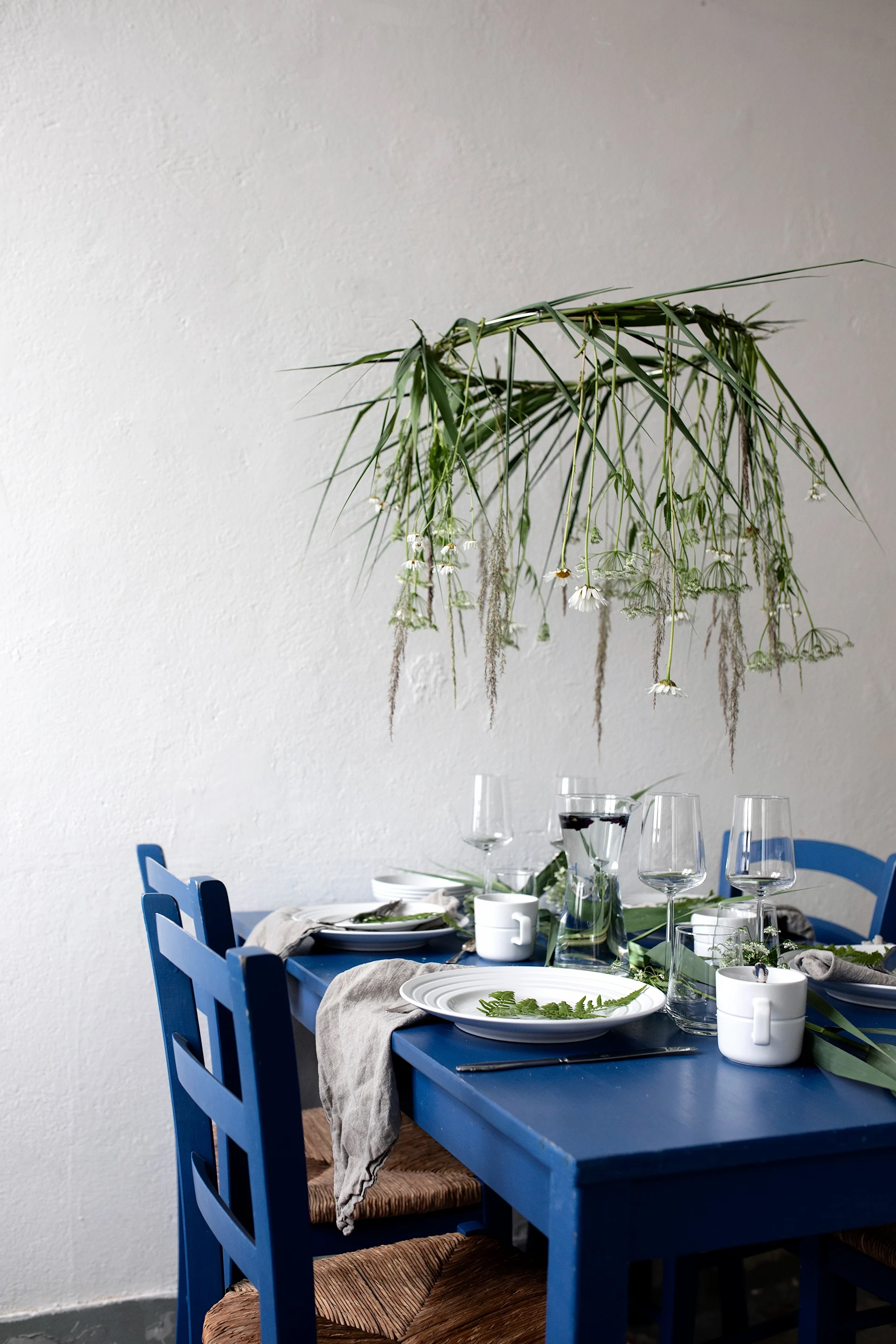 A blue dining table set with the NJRD Lines porcelain set, with white walls with greenery hanging over the table make the perfect Mediterranean themed decor.