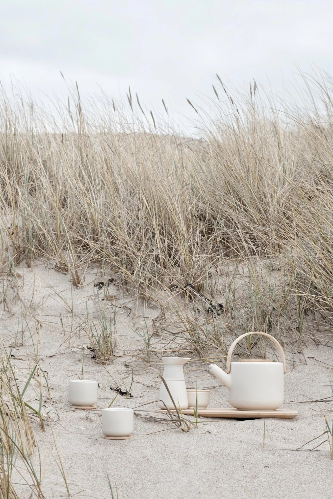 Light-colored ceramic tea set with a teapot, carafe, and cups on a wooden tray, nestled in sand dunes with dry grass.