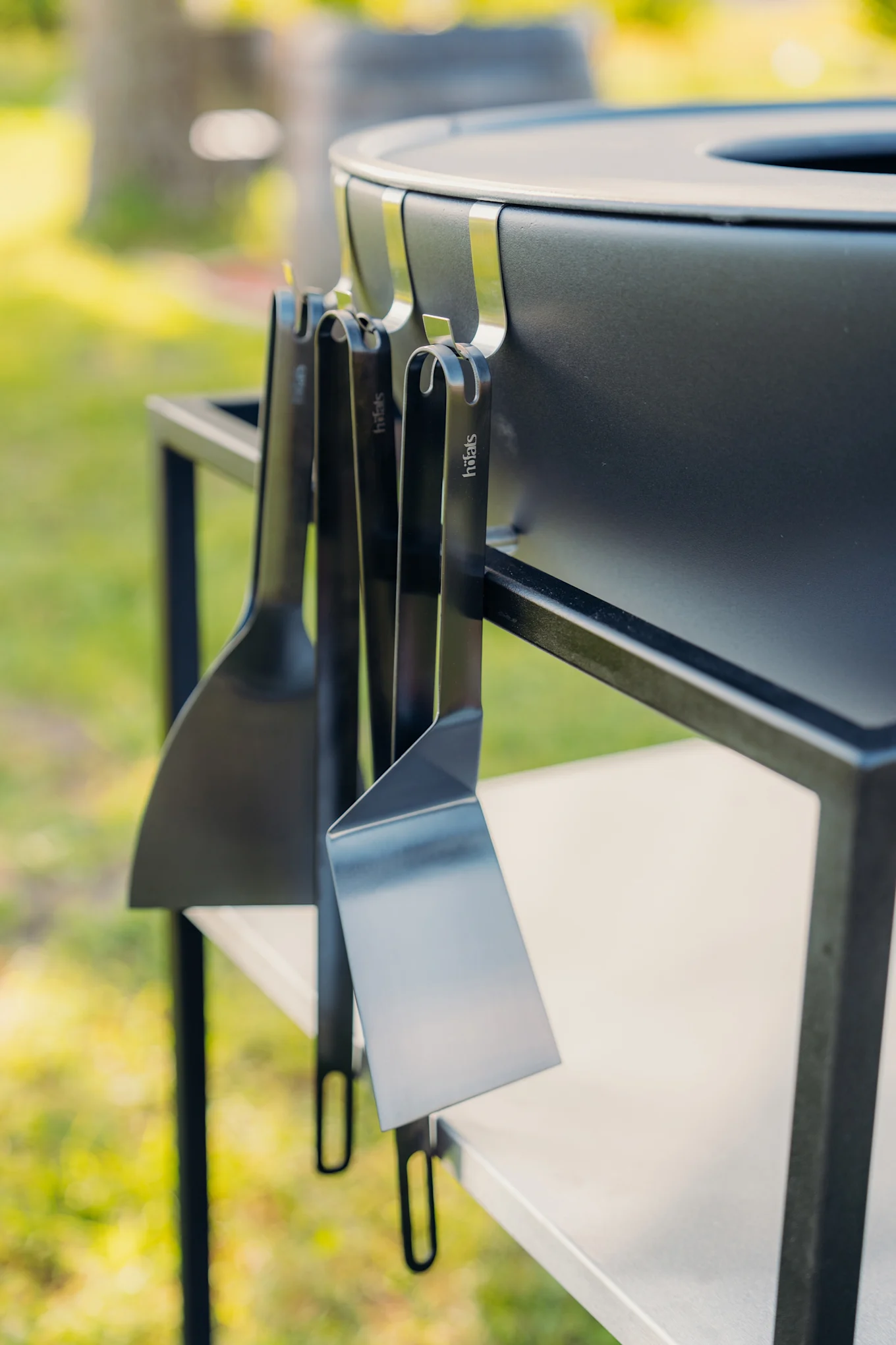 Close-up of a black outdoor grill with three black höfats spatulas hanging from its side in a garden setting.