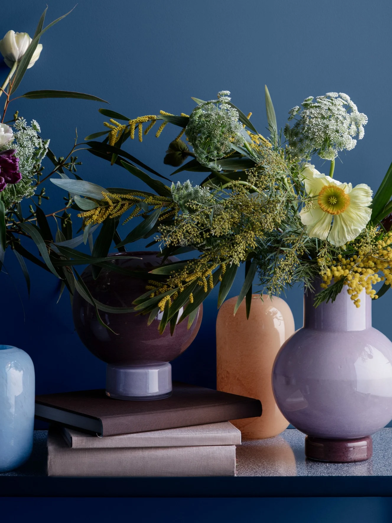 Vibrant floral arrangement: colorful vases with yellow mimosa, white flowers, green leaves, on blue surface, with stacked books.