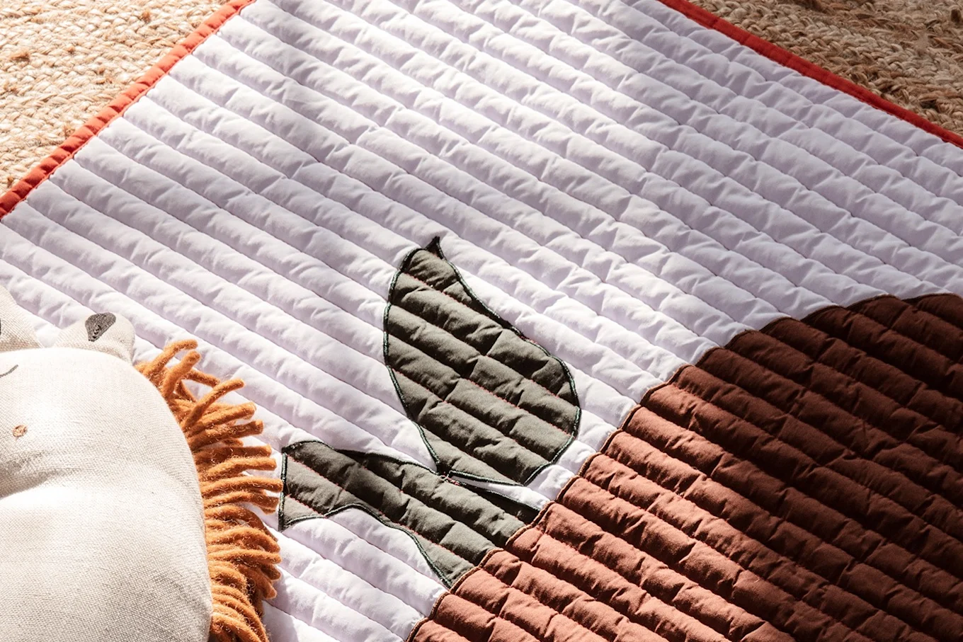Quilted playmat with a lion plush, featuring white, brown, and dark gray leaf patterns on a woven rug.