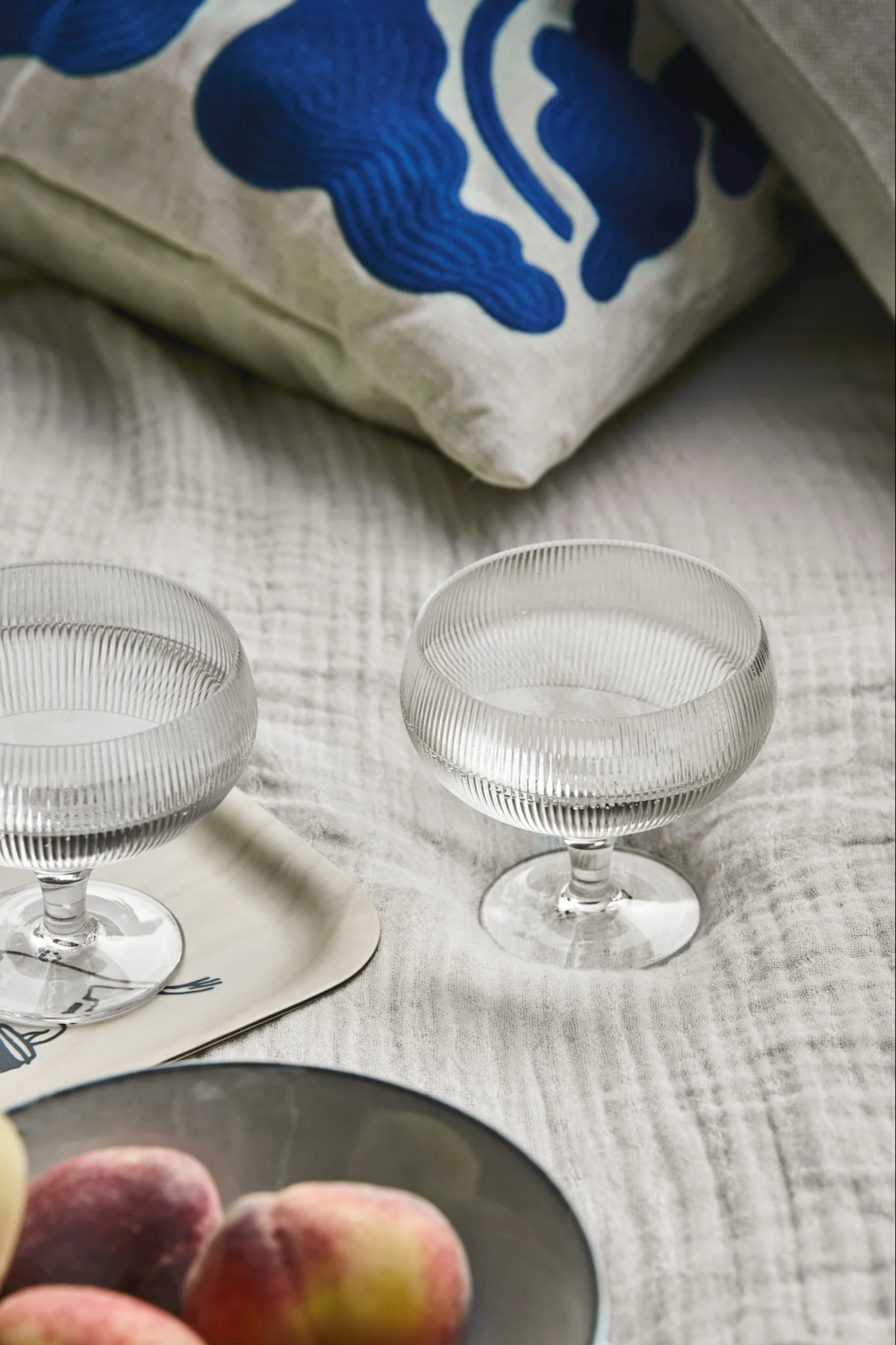 Two ribbed glass coupette glasses on a light tray, with peaches in a bowl and a blue patterned cushion in the background.
