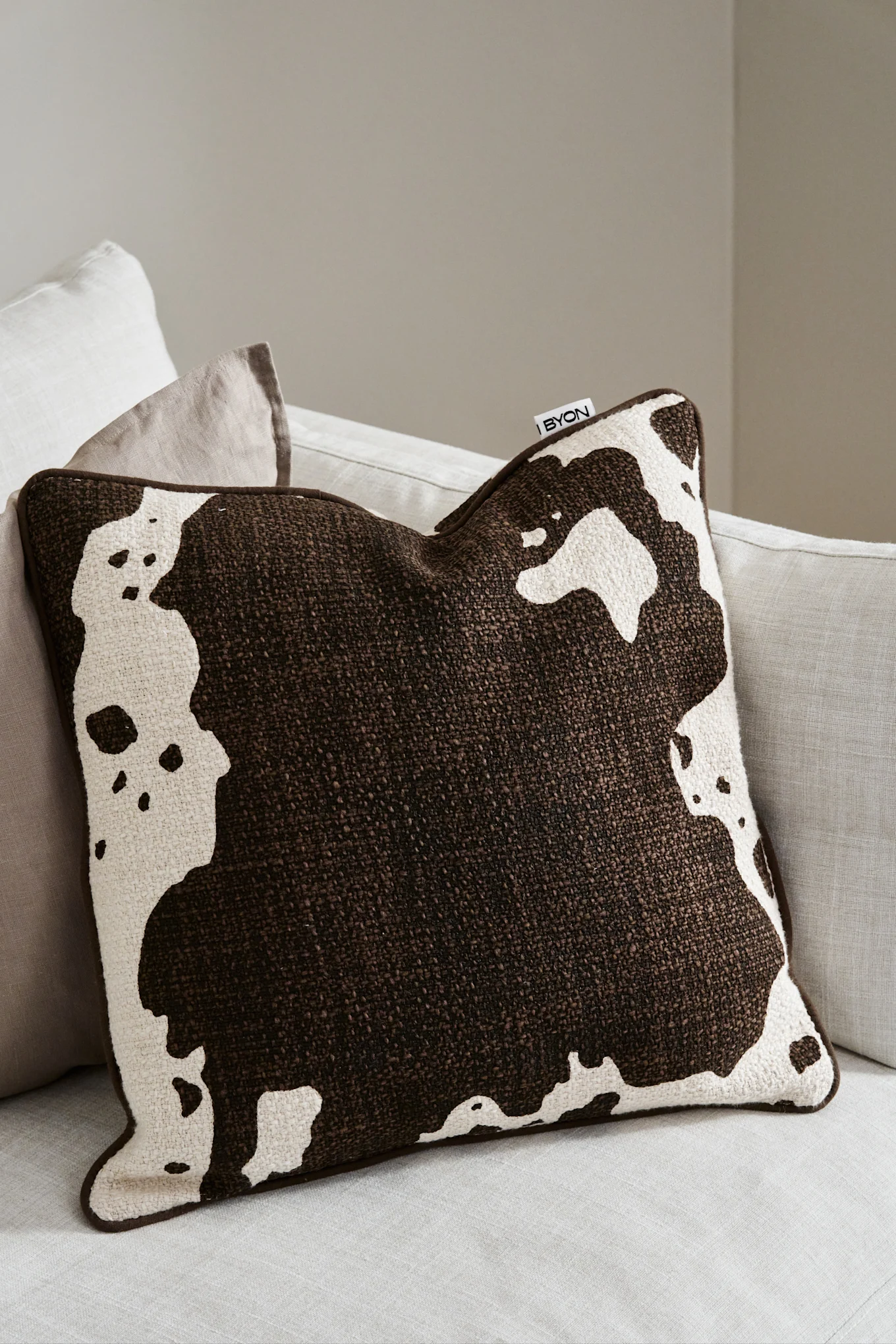 A brown and off-white cow print throw pillow with a textured weave rests on a light-colored sofa.