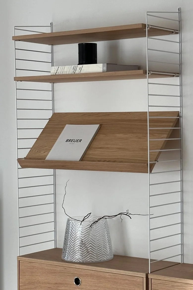 Light wood and white wire shelf unit with books, a black container, a zig-zag glass vase with branches, and wooden drawers.
