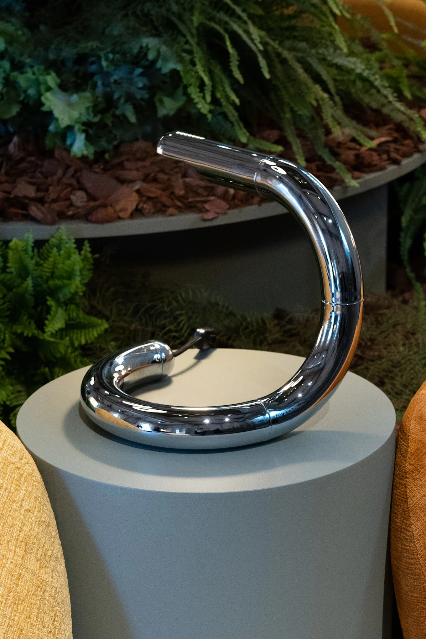 A sleek, curved chrome lamp rests on a grey pedestal, surrounded by green foliage and textured yellow and orange cushions.