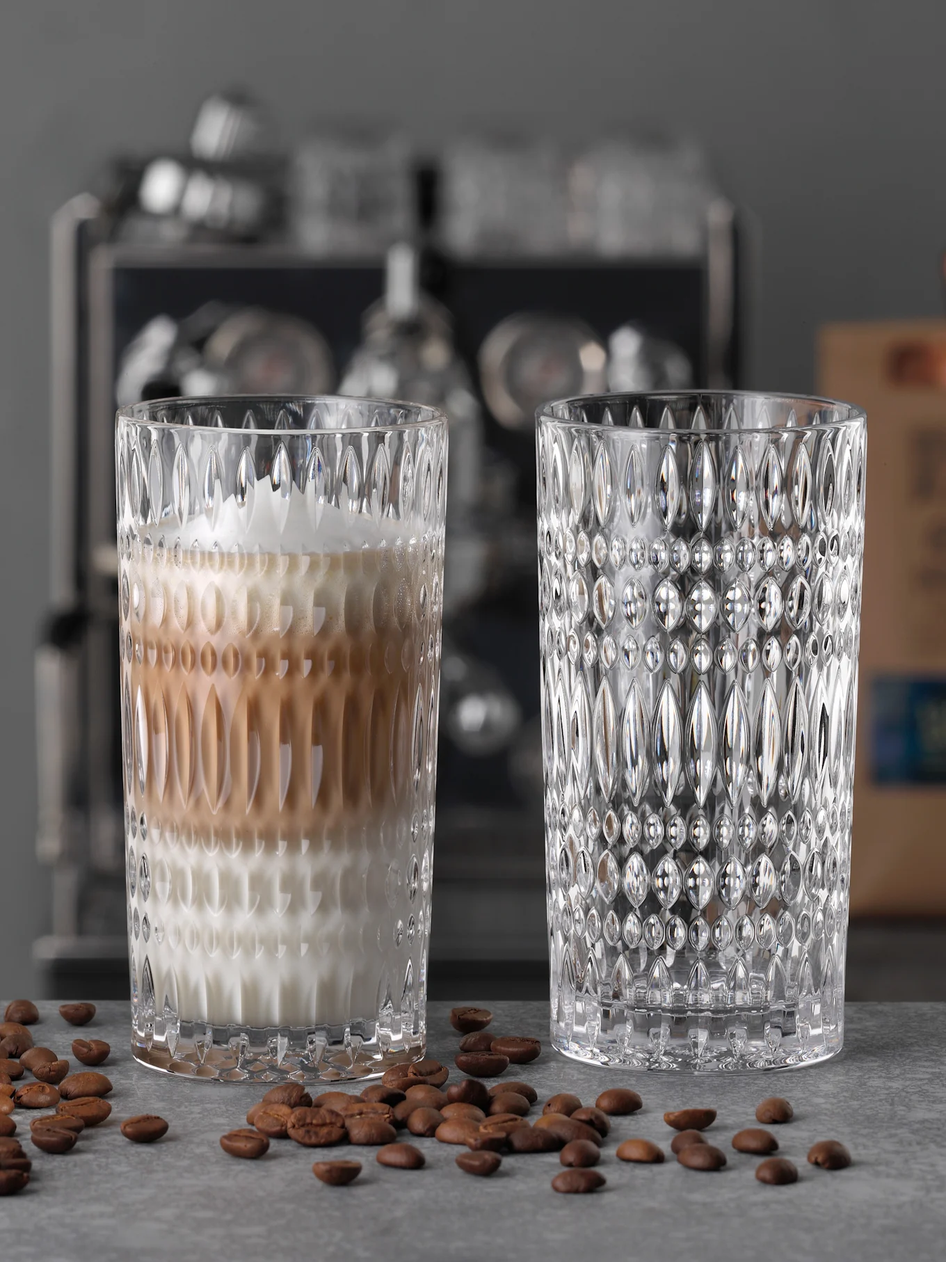 Two ornate clear glass tumblers, one with a layered latte, and an empty one, surrounded by coffee beans on a grey surface.