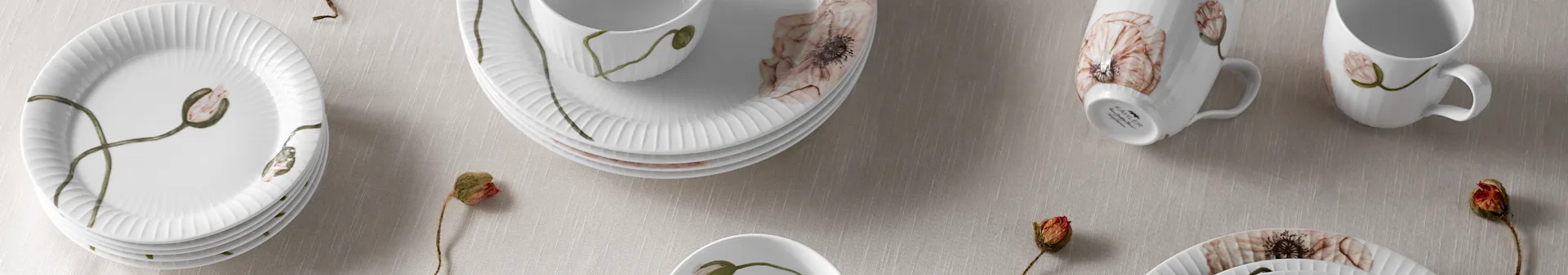 White ribbed porcelain plates, bowls, and mugs with poppy flower designs on a light fabric, with dried poppy pods.
