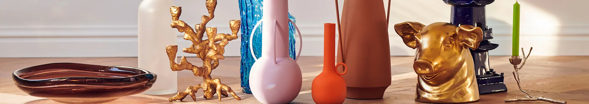 Home decor objects: a brown glass bowl, colorful vases, golden candle holders, and a golden pig sculpture on a wooden floor.