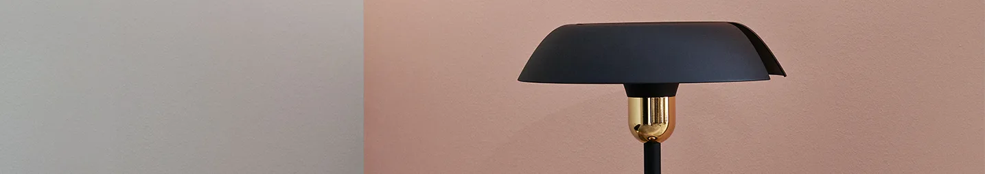 A black lamp with a gold base sits against a two-toned wall, grey on the left and pink on the right.