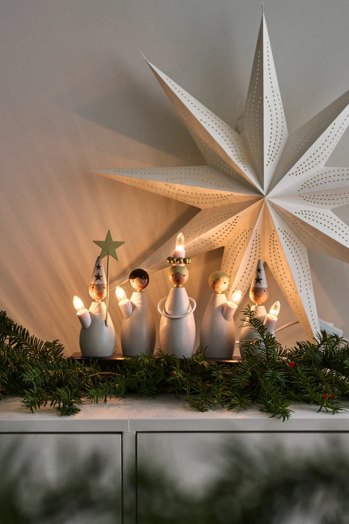Five white wooden angel figures holding candles, with a green garland and a large white star lantern in the background.