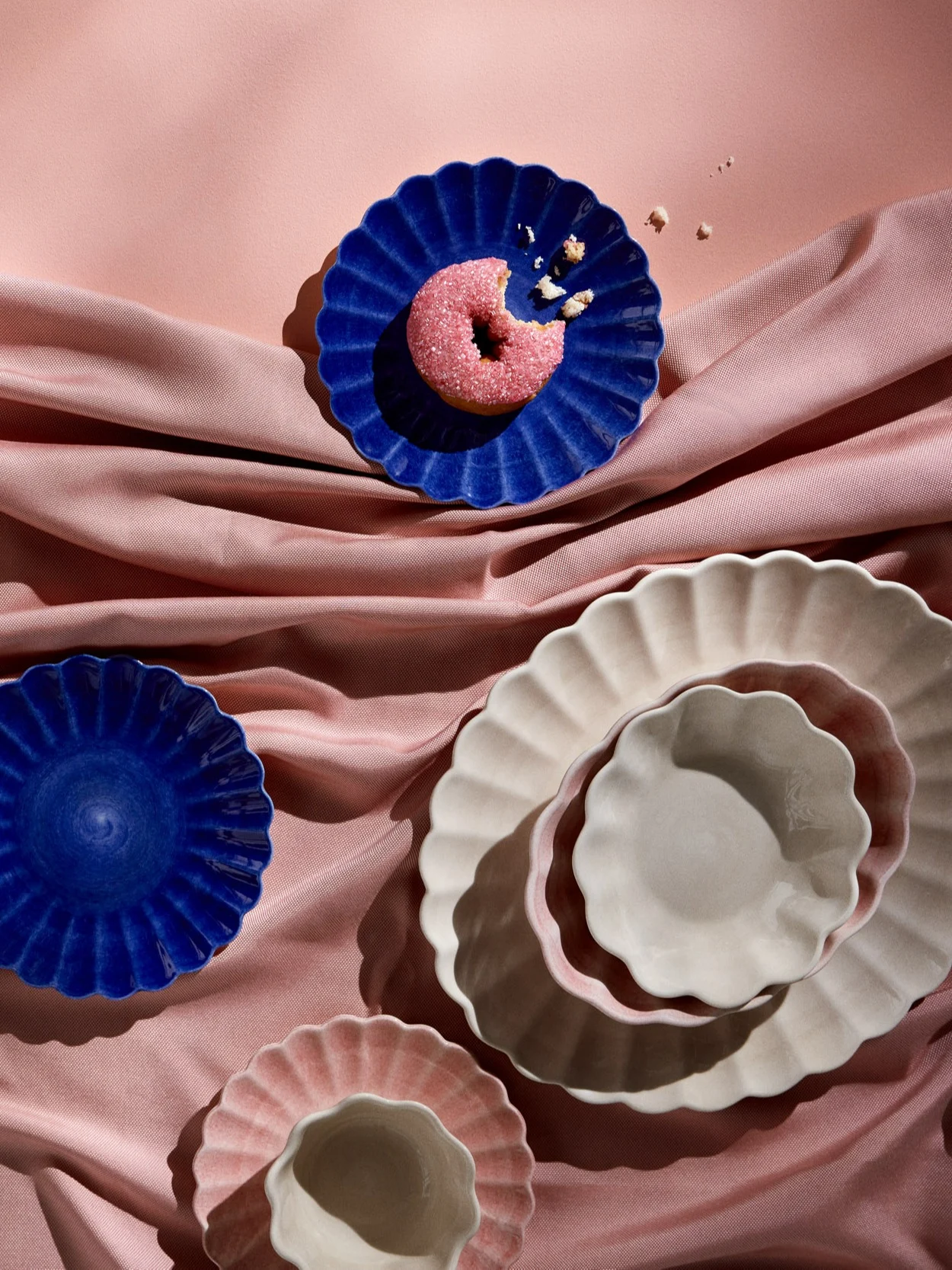 Pink sprinkle donut on a blue scalloped plate, with various blue, pink, and off-white scalloped plates on pink draped fabric.