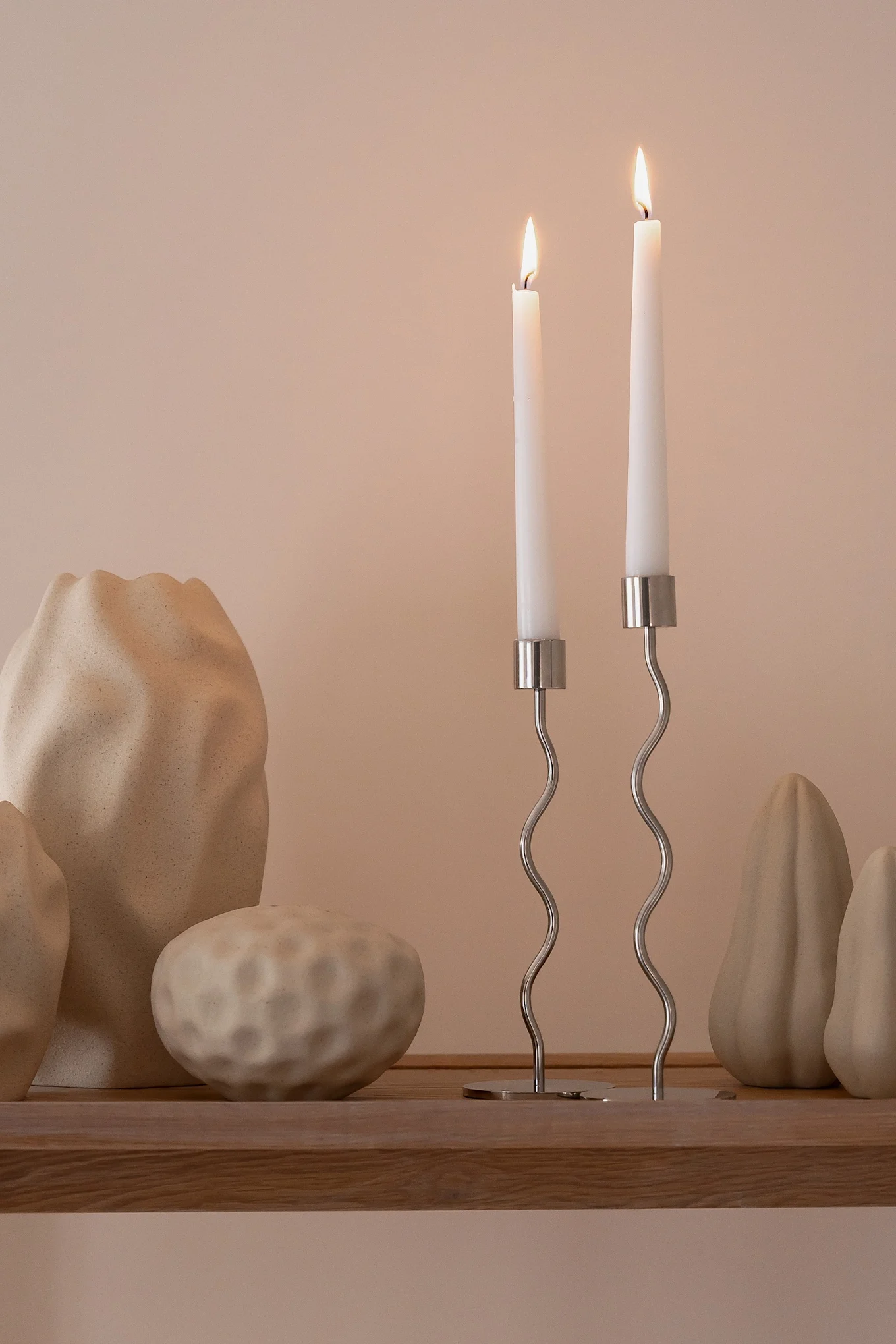 Two lit white taper candles in wavy silver candlesticks on a wooden shelf, with textured beige decorative objects.