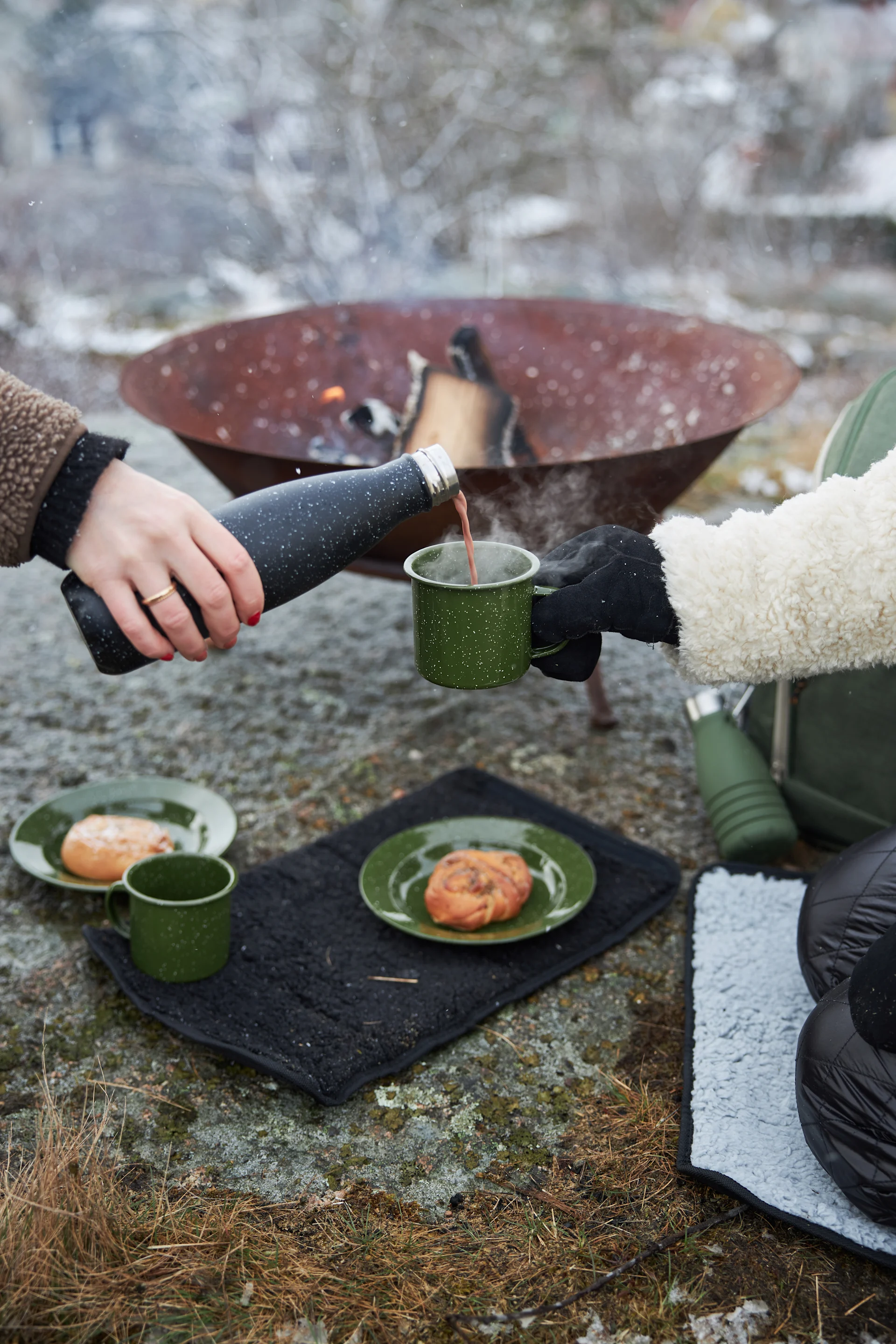 Two people pouring a hot drink from a black thermos into a green mug outdoors, with a fire pit and pastries.
