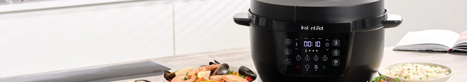 A black multi-cooker cooking seafood paella, with a bowl of rice on the side.