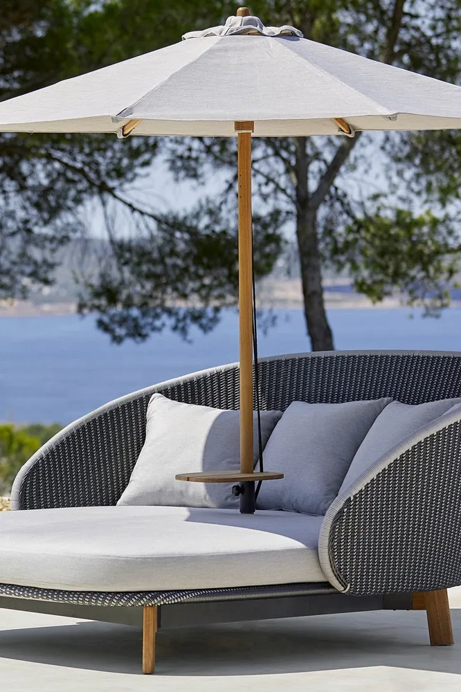 An outdoor lounge chair with light grey cushions and a matching umbrella, set by the sea with trees in the background.