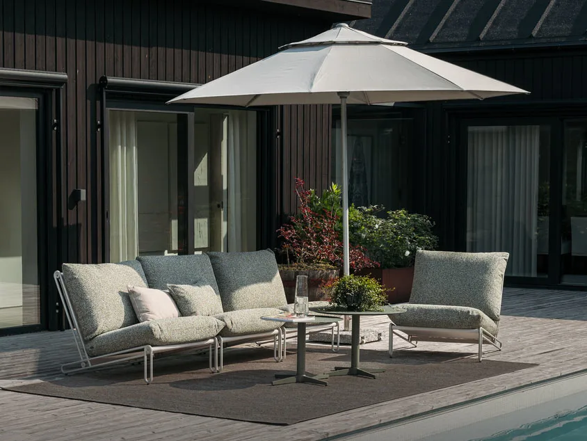 Outdoor patio with a light grey modular sofa, matching armchair, two tables, and a large umbrella on a wooden deck by a pool.