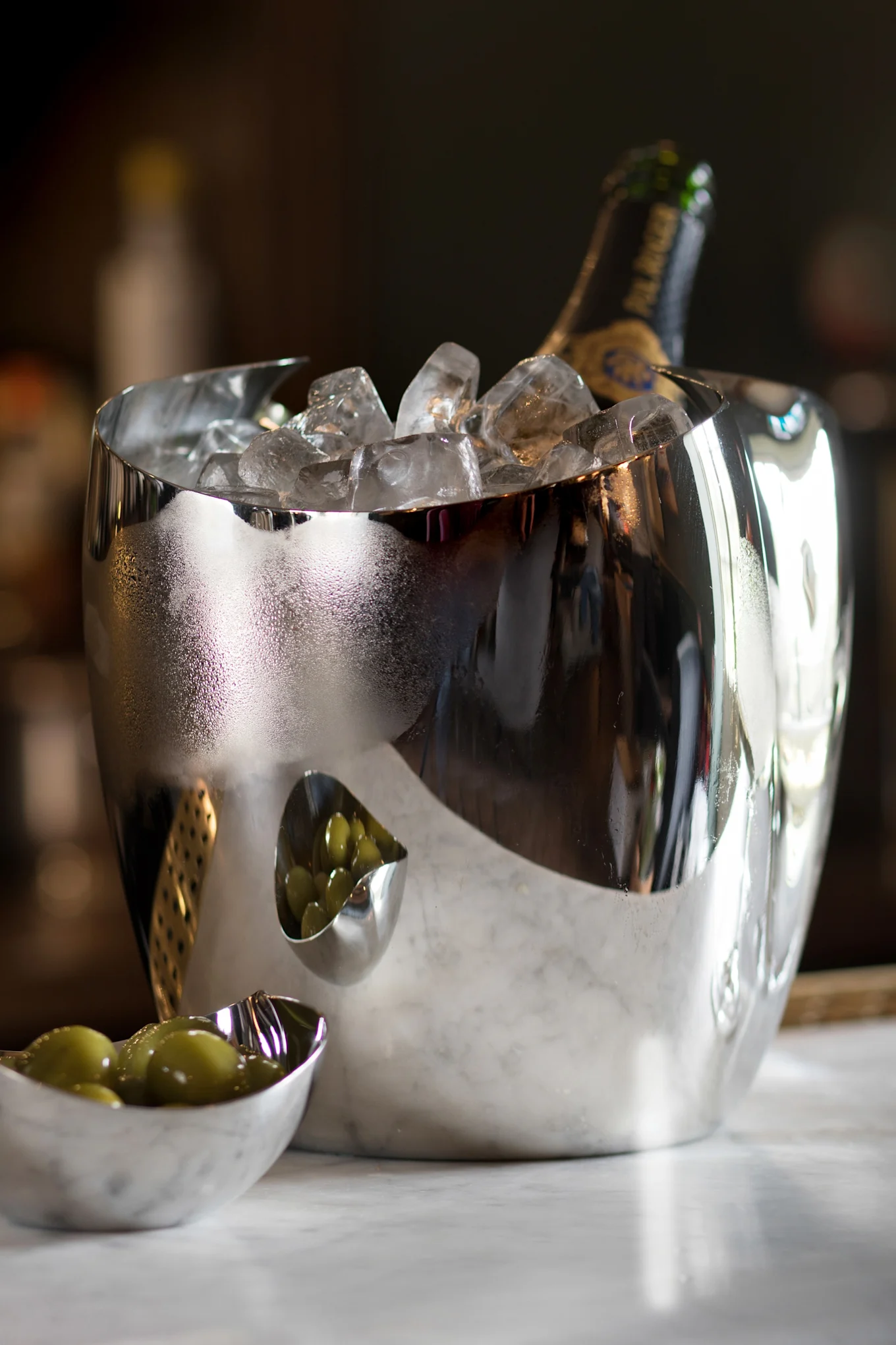 A reflective silver ice bucket with a champagne bottle and ice, next to two small bowls of green olives on marble.
