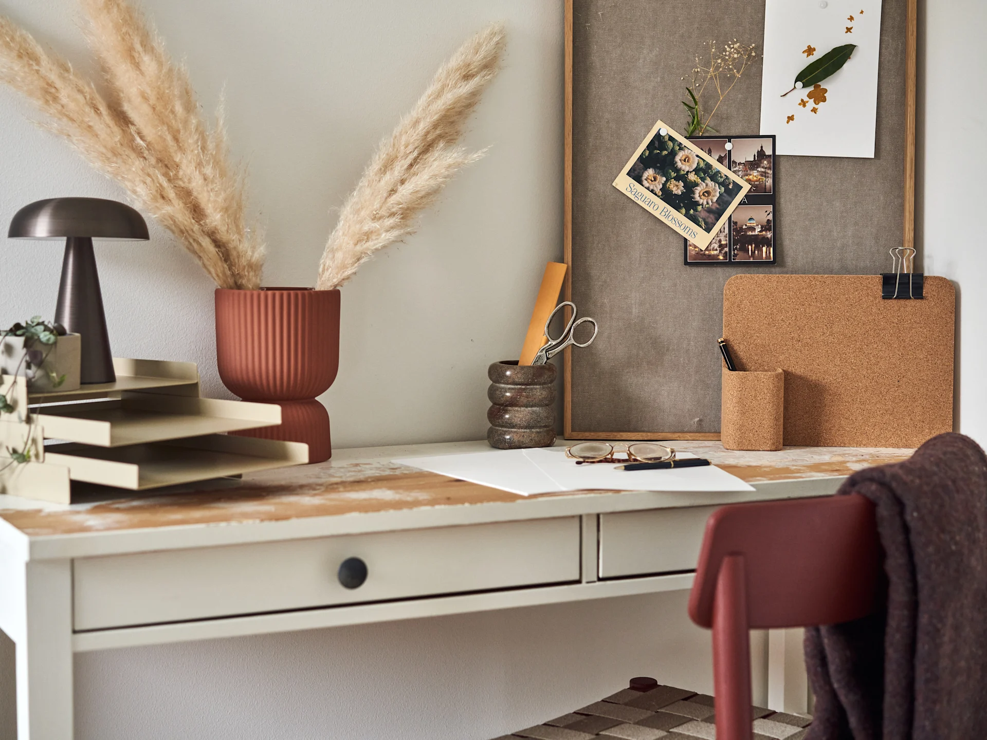 Desk with a dark lamp, terracotta vase with pampas grass, beige letter tray, marbled pen holder, cork board, and a framed bulletin board.
