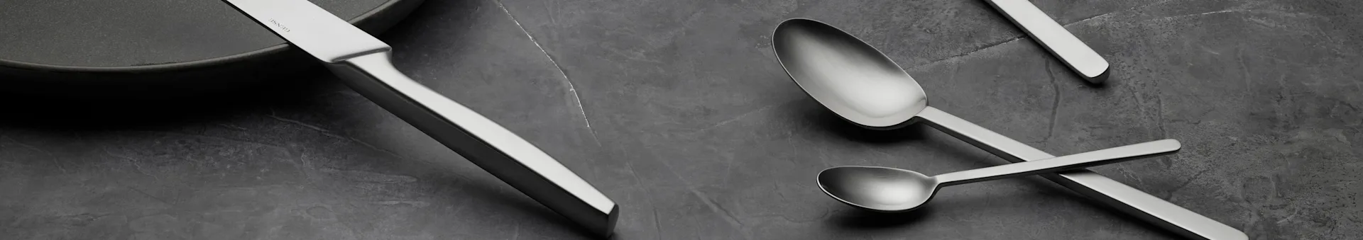 Sleek silver knife and two spoons on a dark, textured surface, with a dark plate visible.