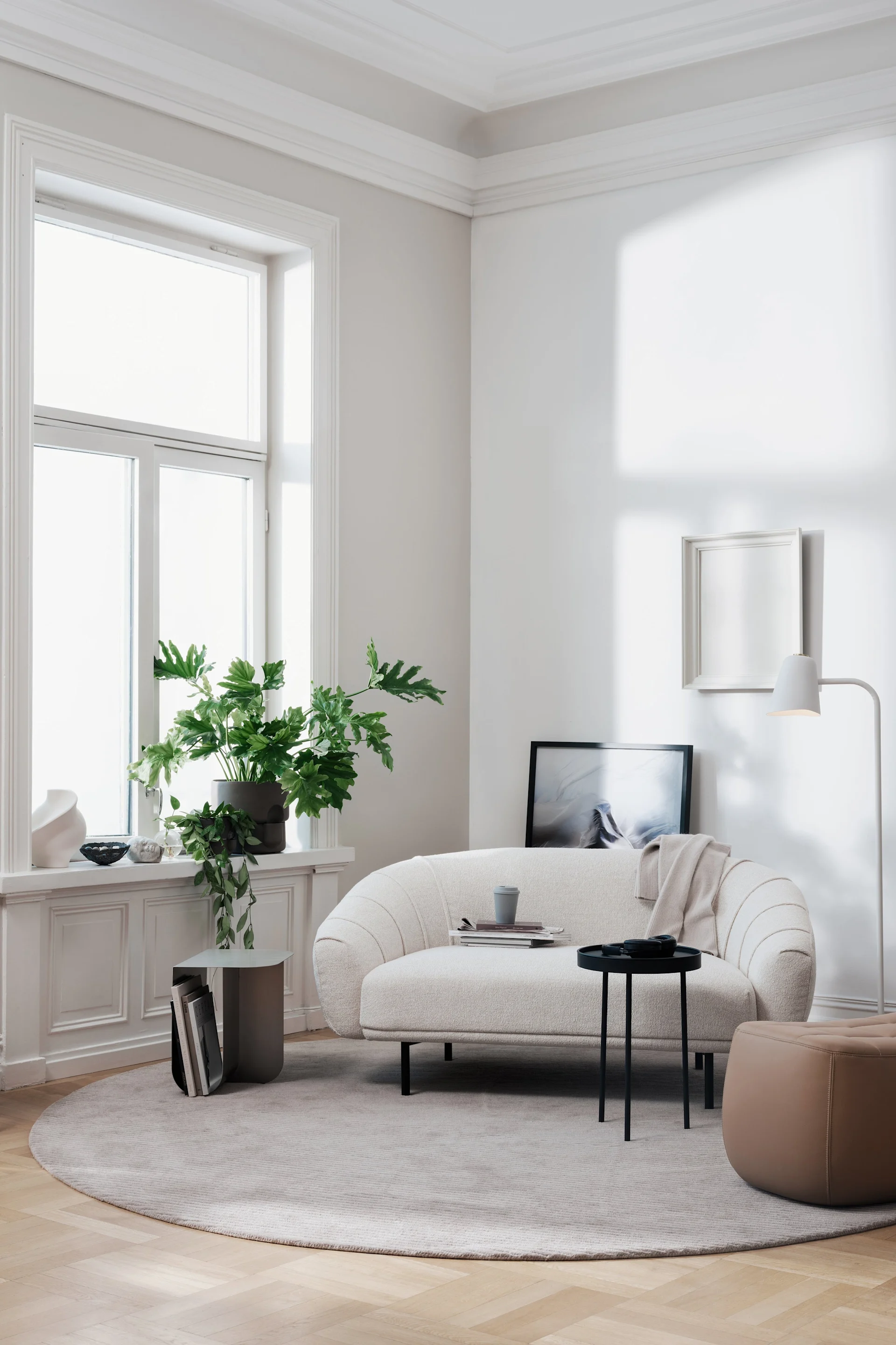 A bright living room with a curved, light beige sofa, a round grey rug on a wooden floor, plants by the window, and a floor lamp.